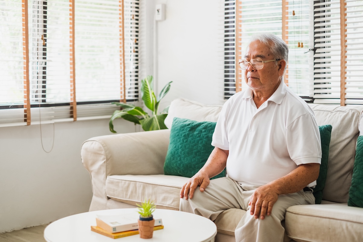 Senior old Asian man sitting on sofa praticing yoga and meditation at home, retirement and wellbeing concept